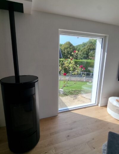 Pièce de vie rénovée avec sol en bois, poêle moderne et grande porte vitrée donnant sur le jardin.
