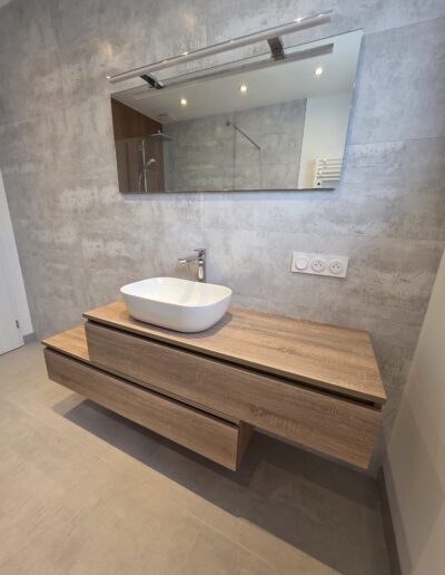 Salle de bain rénovée avec meuble suspendu en bois, vasque blanche et miroir avec éclairage intégré.